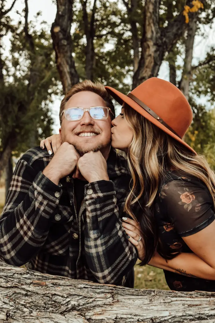 Woman in a rust hat kissing her fiancé's cheek over a log
