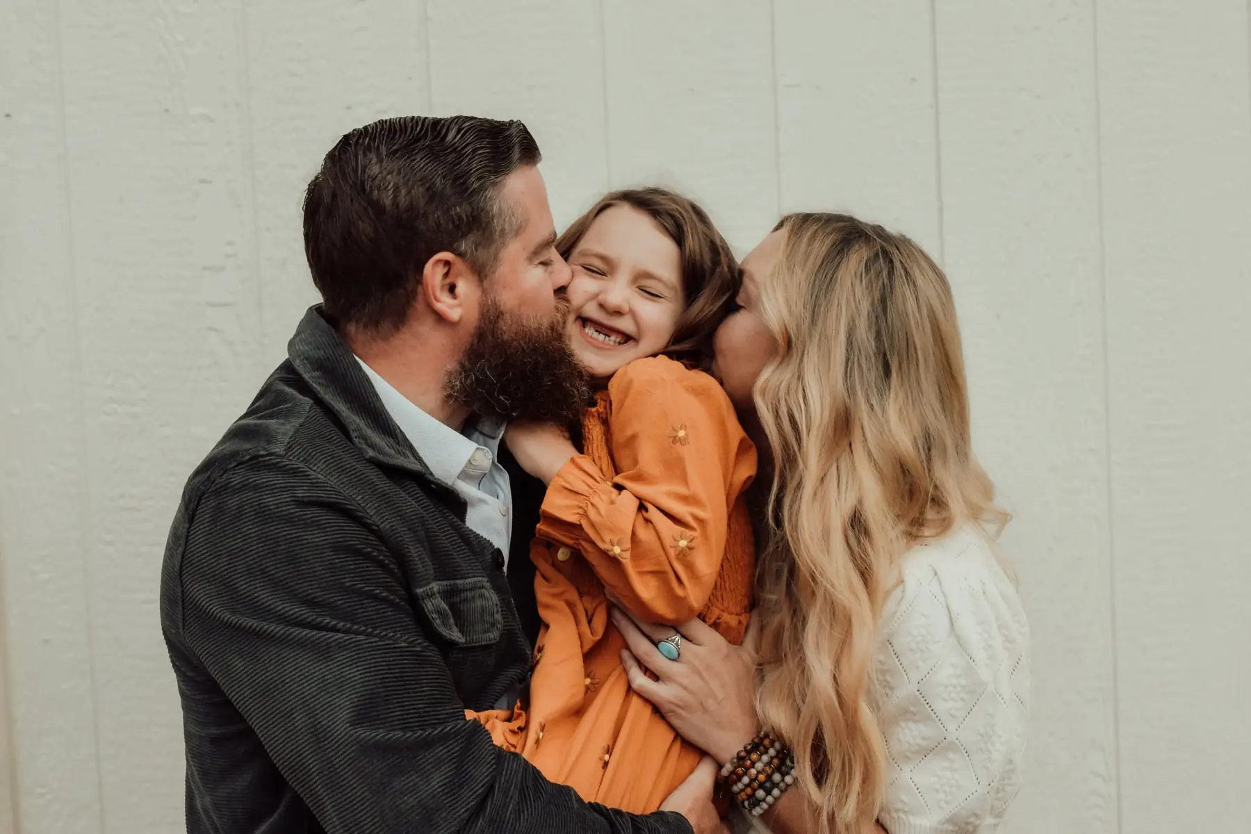 Parents kissing their laughing daughter's cheeks against a cream wall