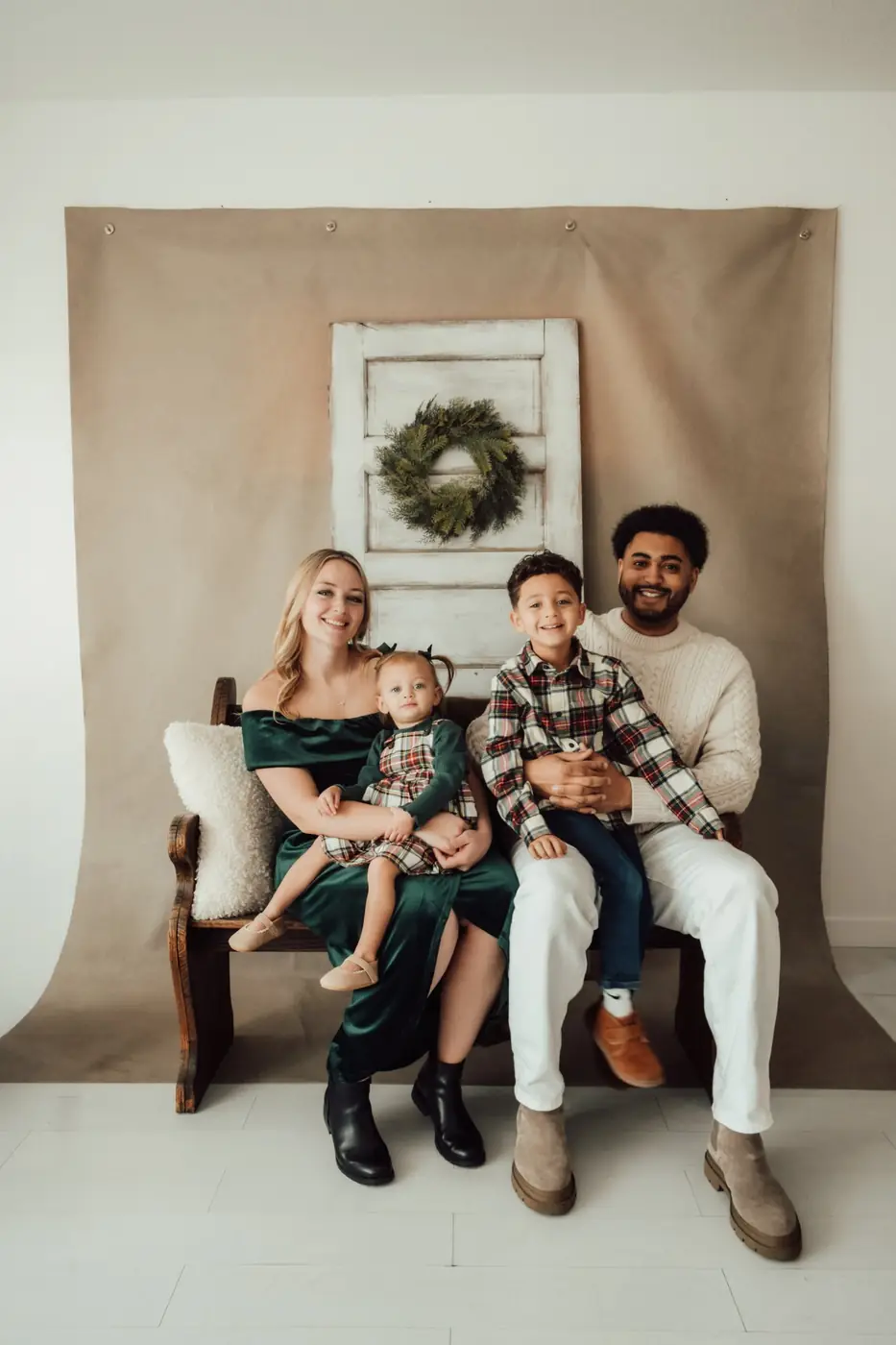 Family in green velvet and plaid seated on a wooden bench with a wreath behind them