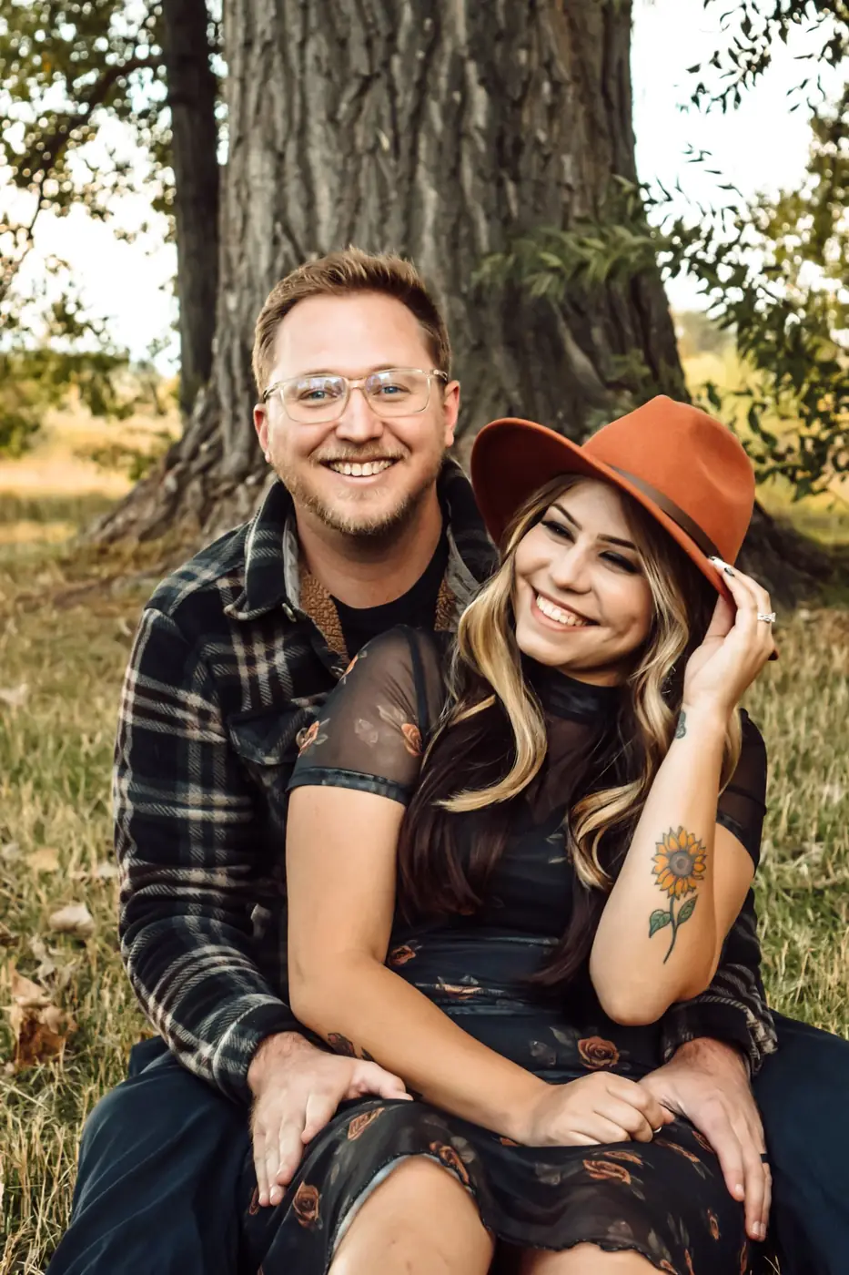 Smiling couple sitting together at the base of a large tree, she tips her rust-colored hat